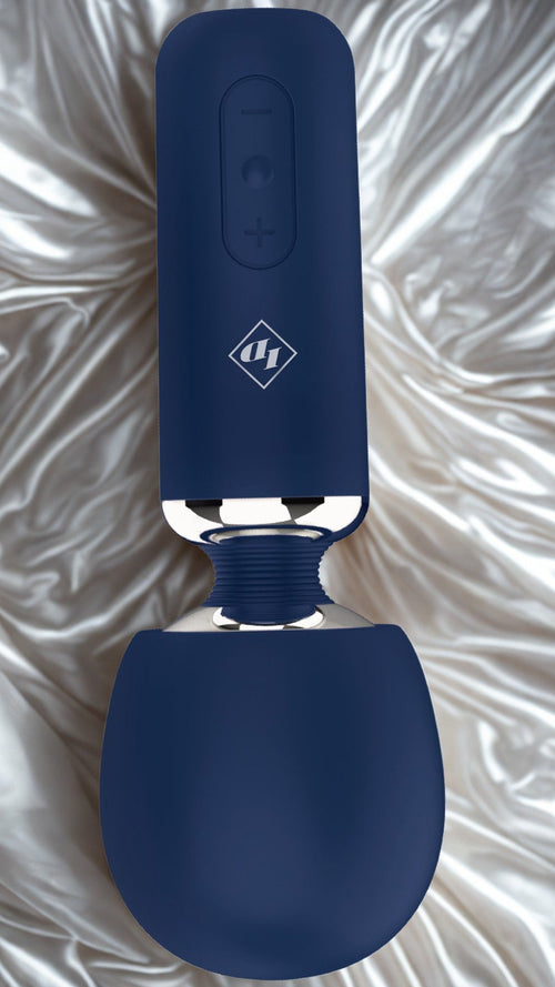 A close-up image of the Give In To Me massager, inverted to show the flexible percussive head, and the simple adaptive control button on white and gold silk sheets. 