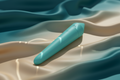 A matte turquoise compact wellness device resting on luxurious, rippling silk that transitions from deep teal to pale gold. Shimmering, aquatic light reflections are dappled across the device and fabric, highlighting its sleek, waterproof design and sensory focus.