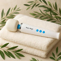 A white Magic Wand Mini wellness device resting on a stack of plush, cream-colored towels. Framed by soft eucalyptus leaves, this scene visualizes an easy grip, gender neutral inclusivity and Simple Elegance. The clean, spa-like environment emphasizes Medical-Grade silicone and Adaptive Sensory Comfort.