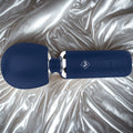 Give Into Me Wellness Wand for Flexible Percussive Care, shown in deep blue on a luxury white and gold brocade bedspread, highlighting its Ergonomic Stability Grip and flexible neck.