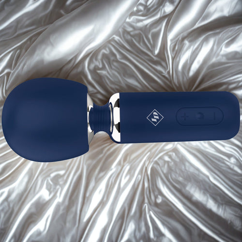 Give Into Me Wellness Wand for Flexible Percussive Care, shown in deep blue on a luxury white and gold brocade bedspread, highlighting its Ergonomic Stability Grip and flexible neck.